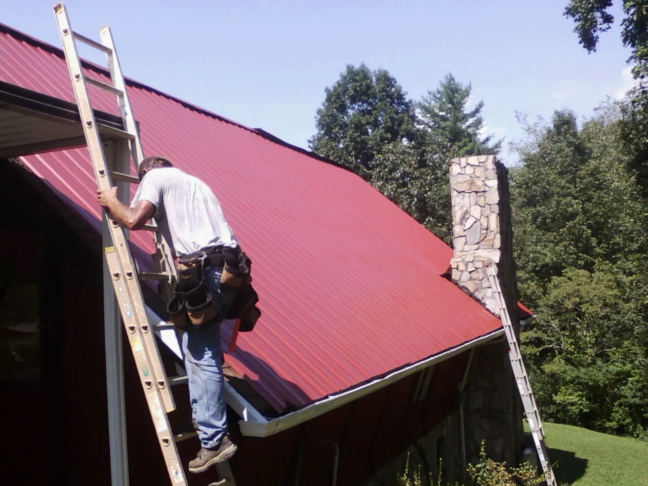 Roofer on a ladder beside a red metal roof and stone chimney — Soffit & Fascia Repair by Roofing Brothers in Cullman